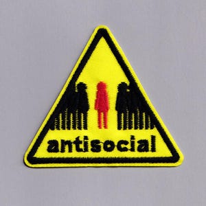 Puede incluir: Parche triangular amarillo con borde negro y la palabra "antisocial" en negro. El diseño presenta tres figuras estilizadas: dos negras y una roja, simbolizando la dinámica social. Un accesorio único.