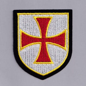 May include: Embroidered patch with a red cross and yellow trim on a white shield. The patch has a black border. The design is a heraldic symbol. The patch is suitable for sewing onto clothing or bags.