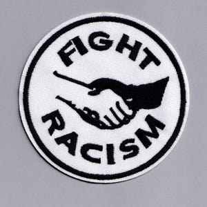 Könnte beinhalten: Runder, weißer Aufnäher mit schwarzem Text und einer schwarzen Grafik von zwei Händen, die sich schütteln. Der Text lautet "FIGHT RACISM" in einem kreisförmigen Muster um die Grafik.