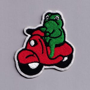 May include: Embroidered patch featuring a green frog riding a red scooter. The frog has large eyes and is centered on the scooter. The scooter has black wheels and a white border. The patch is on a gray background.
