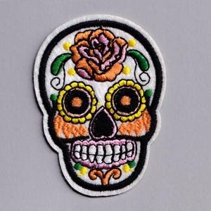 May include: Embroidered skull patch with a white background, black outline, and colorful floral details. The design features a large orange and pink rose, two yellow and orange sunflower-like eyes, and a pink mouth with white teeth.