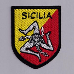 May include: Embroidered patch featuring the Sicilian flag. The patch has a shield shape with a yellow upper left and red lower right background. The design includes the word "SICILIA" and a triskeles symbol with three legs and a face.