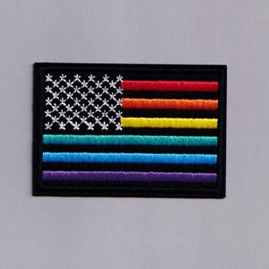May include: Embroidered patch depicting a stylised American flag in rainbow colours. The stars are white, and the stripes are red, orange, yellow, green, blue, and purple. The patch has a black border.