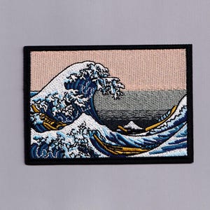 May include: A rectangular embroidered patch featuring a stylized ocean wave in shades of blue, white, and gold, set against a pale pink sky and a dark gray sea. The design is framed by a thick black border with detailed stitching throughout.