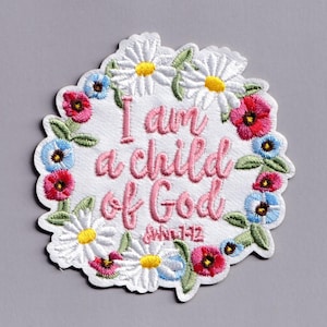 May include: Embroidered patch with the text "I am a child of God" surrounded by a floral wreath. The patch features white daisies with yellow centres, pink, red, and blue flowers, and green leaves. The text is in pink.