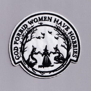 Pode incluir: Patch bordado com o texto "GOD FORBID WOMEN HAVE HOBBIES" em um design circular. O patch apresenta uma ilustração em preto e branco de três figuras com chapéus de bruxa de mãos dadas, cercadas por árvores e pássaros.