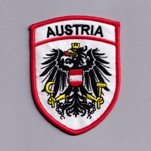 May include: A shield-shaped embroidered patch featuring the Austrian coat of arms with a black eagle, a red and white shield on its chest, a yellow sickle, and a yellow hammer. The word AUSTRIA is stitched in black on a white background with a red border.