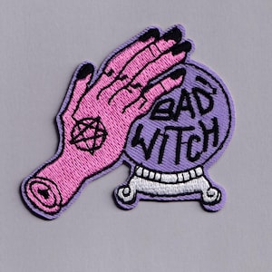 Könnte beinhalten: Gestickter Aufnäher mit einer rosa Hand mit schwarzen Fingernägeln und einem Pentagramm, die eine lila Kristallkugel hält. Die Kristallkugel trägt den schwarzen Schriftzug "BAD WITCH". Der Aufnäher hat einen hellvioletten Rand.