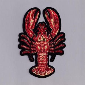 May include: Embroidered patch featuring a detailed red lobster design with black outlining. The lobster has textured claws and body, with accents of gold thread. This patch is suitable for jackets, bags, or other fabric items.