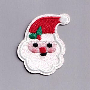 May include: Embroidered Santa Claus patch. The patch features a red hat, white beard, and pink face. It has a green holly accent and black eyes. The edges are outlined in white thread, creating a festive holiday design.