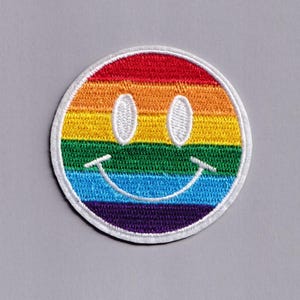 Puede incluir: Parche bordado con un diseño de cara sonriente arcoíris. El parche presenta un borde blanco y una cara sonriente con ojos ovalados. El arcoíris consta de rayas rojas, naranjas, amarillas, verdes, azules y moradas.