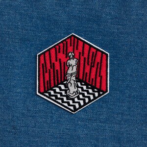 May include: A red and black embroidered patch featuring a white silhouette of a person standing in a room with a black and white checkered floor. The text "C.A.C.H.E.R." is embroidered in black on the red walls.