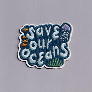 Pu&ograve; includere: Toppa ricamata con la scritta "Save Our Oceans" in azzurro, bordata di bianco, su sfondo blu scuro. La toppa presenta illustrazioni di pesci, una medusa e alghe.