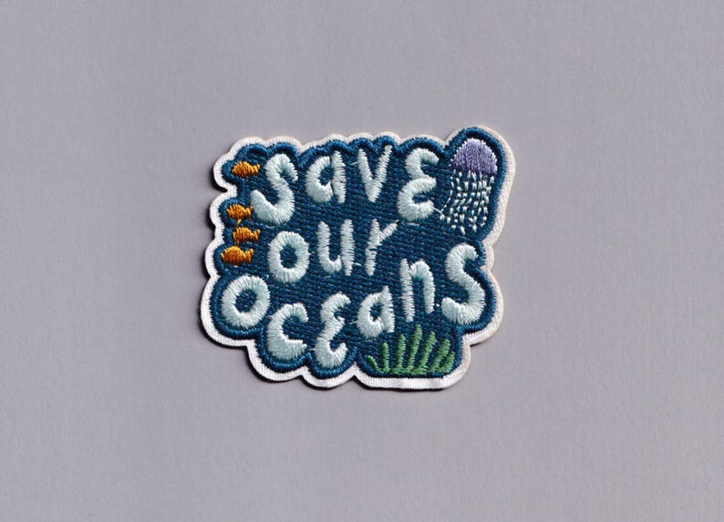 Pu&ograve; includere: Toppa ricamata con la scritta "Save Our Oceans" in azzurro, bordata di bianco, su sfondo blu scuro. La toppa presenta illustrazioni di pesci, una medusa e alghe.