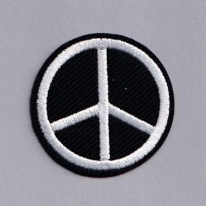 May include: Circular black patch featuring a white peace symbol. The peace sign is a white circle with three lines pointing downwards. The patch has a white border. The background is a light grey colour.