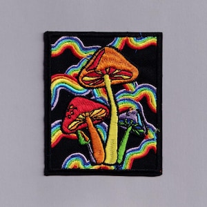Op de afbeelding: Geborduurde patch met drie kleurrijke paddenstoelen en regenboogaccenten op een zwarte achtergrond. De patch is rechthoekig met een zwarte rand. De paddenstoelen hebben oranje, rode en paarse hoeden.