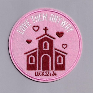 Pode incluir: Um patch bordado redondo rosa com uma igreja bordô e uma cruz. O texto "LOVE THEM ANYWAY" está acima da igreja, com três corações vermelhos. O texto "LUCK 23:34" está abaixo.