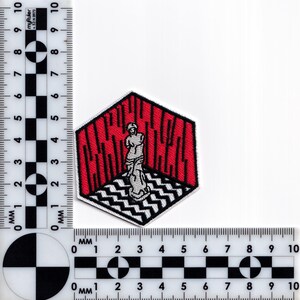 May include: An embroidered patch featuring a red and white geometric design. A white figure stands in the center of the design, which is surrounded by a black outline.