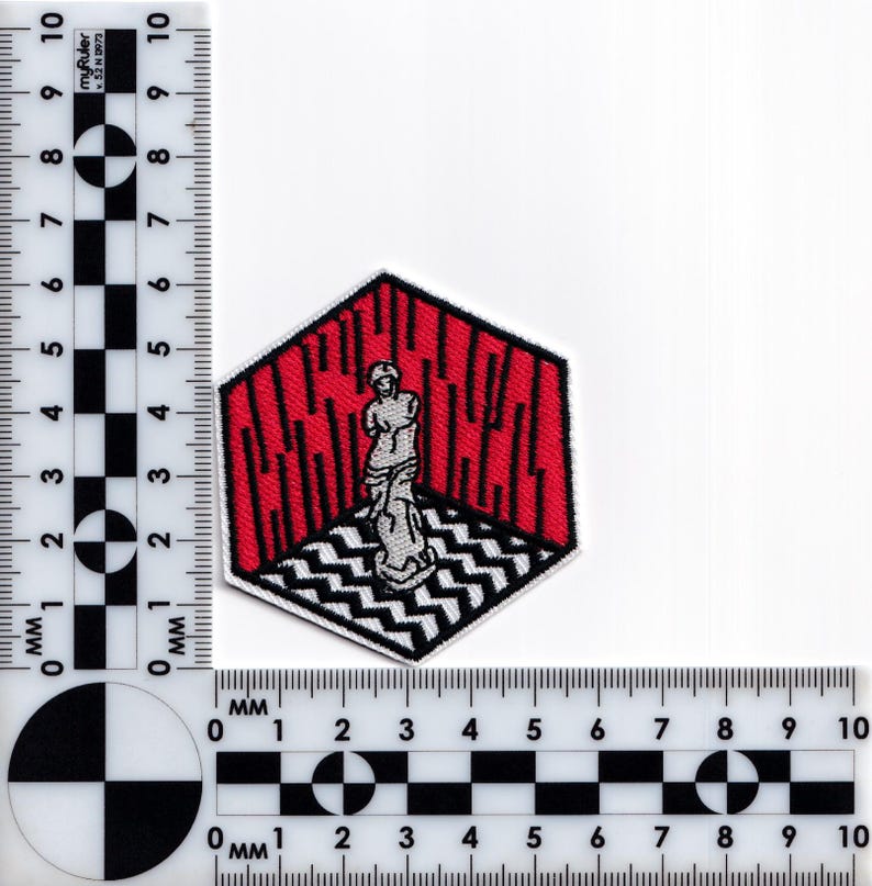 May include: An embroidered patch featuring a red and white geometric design. A white figure stands in the center of the design, which is surrounded by a black outline.