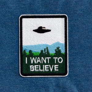 May include: A white and green embroidered patch with a black UFO flying over a green forest and the text "I WANT TO BELIEVE".