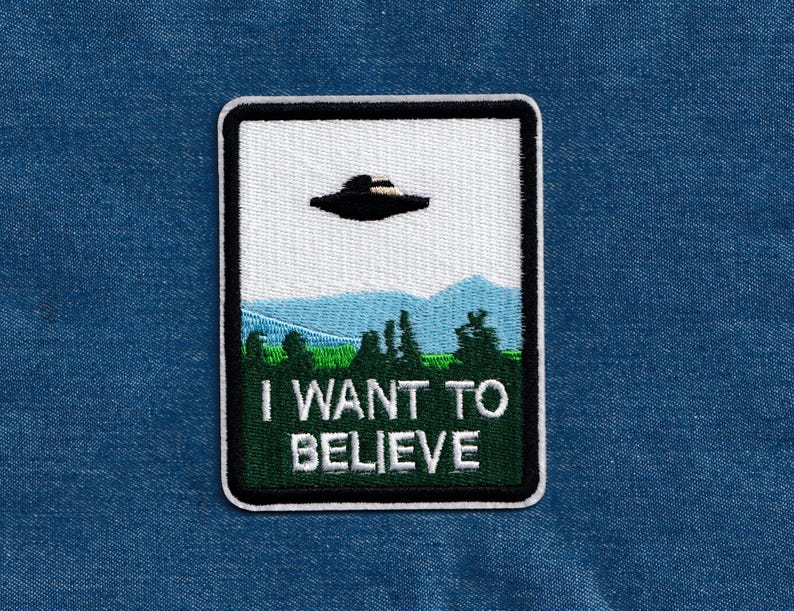 May include: A white and green embroidered patch with a black UFO flying over a green forest and the text "I WANT TO BELIEVE".