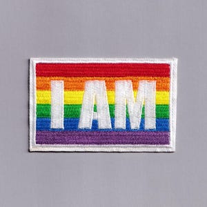 May include: Embroidered rectangular patch with a rainbow design and the text "I AM". The patch features a red, orange, yellow, green, blue, and purple colour scheme, with a white border.