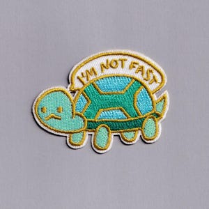 Può includere: Toppa ricamata raffigurante una tartaruga dei cartoni animati con corpo turchese e guscio blu e verde. La tartaruga ha un'espressione triste e una nuvoletta con la scritta "I'M NOT FAST" in lettere dorate. La toppa ha un bordo bianco.