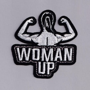 May include: Embroidered patch with a flexing figure and the text "WOMAN UP". The design features a black border with white stitching, and the figure and text are in white. A strong, empowering design.