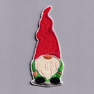 Könnte beinhalten: Gestickter Aufnäher eines Gartenzwergs. Der Zwerg hat eine hohe rote Mütze, einen langen weißen Bart und grüne Kleidung. Der Aufnäher ist mit weißen Nähten umrandet und hat schwarze Schuhe. Der Aufnäher ist zum Aufbügeln bestimmt.