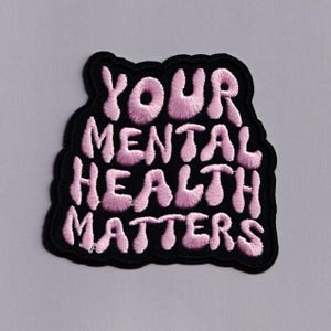 May include: Embroidered patch featuring the text "YOUR MENTAL HEALTH MATTERS" in pink, rounded letters. The text is set against a black, wavy-edged background. The patch is designed to be attached to fabric items.