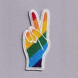 Puede incluir: Parche bordado con forma de mano haciendo el signo de la paz. El parche presenta un diseño de arcoíris con rayas rojas, naranjas, amarillas, verdes, azules y moradas. Los bordes están delineados con hilo blanco.