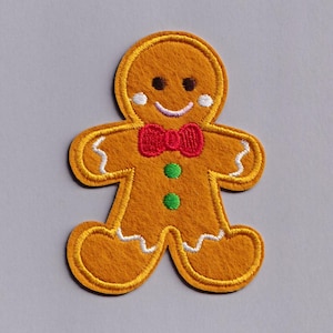 May include: Embroidered gingerbread man patch with a smiling face, red bow tie, and green buttons. The patch has a yellow outline and white accents. Perfect for holiday crafts and apparel.