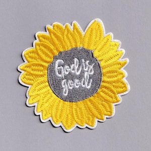 Puede incluir: Parche bordado con un girasol amarillo con un centro gris. El texto "God is good" está bordado en escritura cursiva blanca dentro del centro de la flor. El parche tiene un borde blanco.
