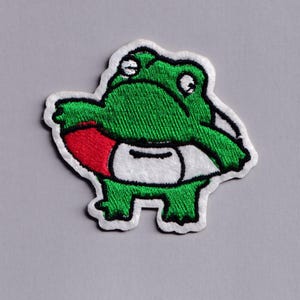 Può includere: Toppa ricamata raffigurante una rana dei cartoni animati verde con un salvagente rosso e dettagli bianchi. La rana ha la pancia bianca ed è bordata di nero. Su sfondo grigio.