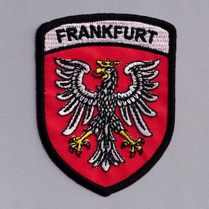 Puede incluir: Un parche bordado en forma de escudo con fondo rojo y un emblema de águila en blanco y negro. El águila tiene garras y una corona amarillas. Sobre el águila, la palabra FRANKFURT está bordada en letras negras sobre una banda blanca.