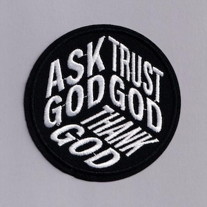 May include: A circular, black embroidered patch featuring the words "ASK GOD TRUST GOD THANK GOD" in white, bold lettering. The patch is designed for application on clothing or accessories, with a simple, clear aesthetic.