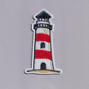 May include: Embroidered patch of a lighthouse with red and white stripes, a black top, and a beige base. The lighthouse has two small arched windows. The patch is outlined in white.