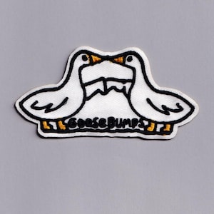 Puede incluir: Parche bordado con dos gansos blancos enfrentados, con contornos negros y picos y patas amarillas. El parche tiene la inscripción "Goosebumps" en letras negras con detalles amarillos.