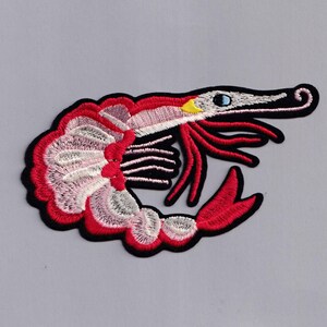 May include: An embroidered iron-on patch of a shrimp, featuring red and white colours. The shrimp is curled up with its tail tucked under its body. The shrimp has a black outline and yellow eyes.