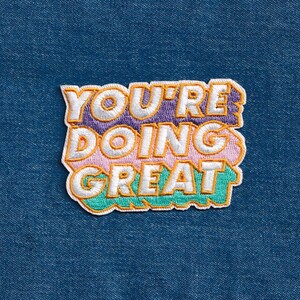 May include: A colorful embroidered patch with the text "You're doing great" in a retro font style. The patch is made of fabric and has a white outline.