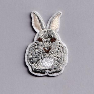 May include: Embroidered patch of a grey and white rabbit with long ears and brown eyes. The bunny patch has a white border and is set against a grey background. Suitable for clothing or accessories.