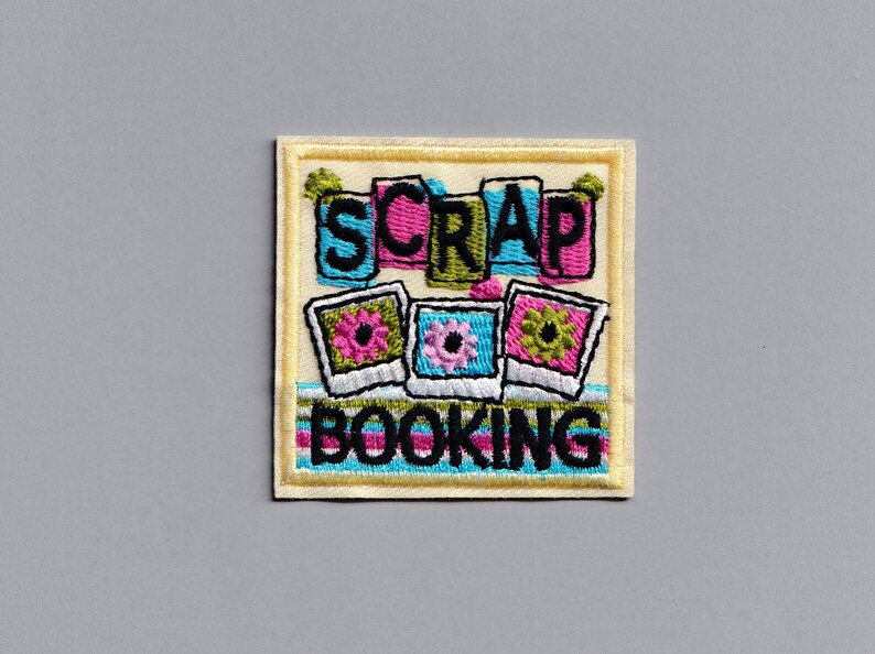 May include: A yellow embroidered patch with the words "Scrap Booking" in black lettering. The patch features three colorful photo frames with flowers.