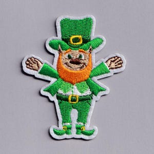 Puede incluir: Parche bordado con un duende vestido de verde, con un sombrero a juego. El duende tiene barba naranja y los brazos extendidos. El parche tiene un borde blanco y está sobre un fondo gris. Ideal para el Día de San Patricio.