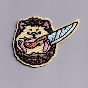 May include: Embroidered patch featuring a cartoon hedgehog holding a knife. The hedgehog has brown quills, pink ears and feet, and a light yellow belly. The knife has a blue blade and a white handle. The patch has a black border.