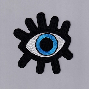 May include: Black embroidered patch with a stylized eye design. The eye has a blue iris and white detailing, set within a black, sunburst-shaped border. Suitable for application on clothing or accessories.