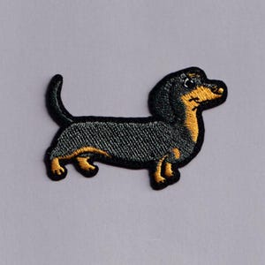 Könnte beinhalten: Gestickter Aufnäher eines Dackelhundes im Profil. Der Hund hat einen schwarzen Körper mit braunen und gelben Akzenten an Beinen, Brust und Gesicht. Für Kleidung oder Accessoires.