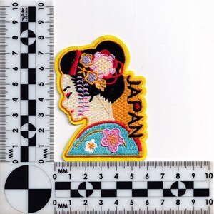 May include: Embroidered iron-on patch featuring a geisha with a floral headdress and the word "JAPAN" in yellow lettering.