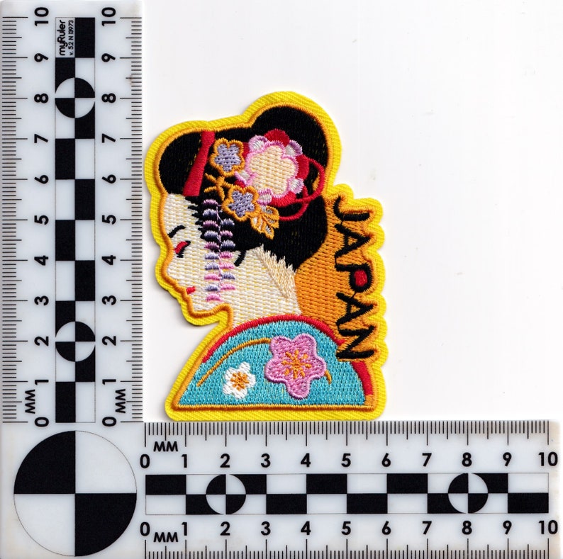 May include: Embroidered iron-on patch featuring a geisha with a floral headdress and the word "JAPAN" in yellow lettering.