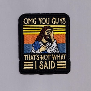 Könnte beinhalten: Gestickter Aufnäher mit schwarzem Rand. Er zeigt den Text "OMG YOU GUYS" über einem Bild von Jesus mit der Hand vor dem Gesicht. Darunter steht der Text "THAT'S NOT WHAT I SAID."