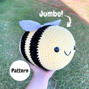 Op de afbeelding: Een grote, gehaakte bijenknuffel met gele en zwarte strepen. De bij heeft zwarte ogen, een lachende mond en kleine grijze vleugels. Het woord "Jumbo!" staat boven de bij en het woord "Pattern" staat in een witte cirkel.